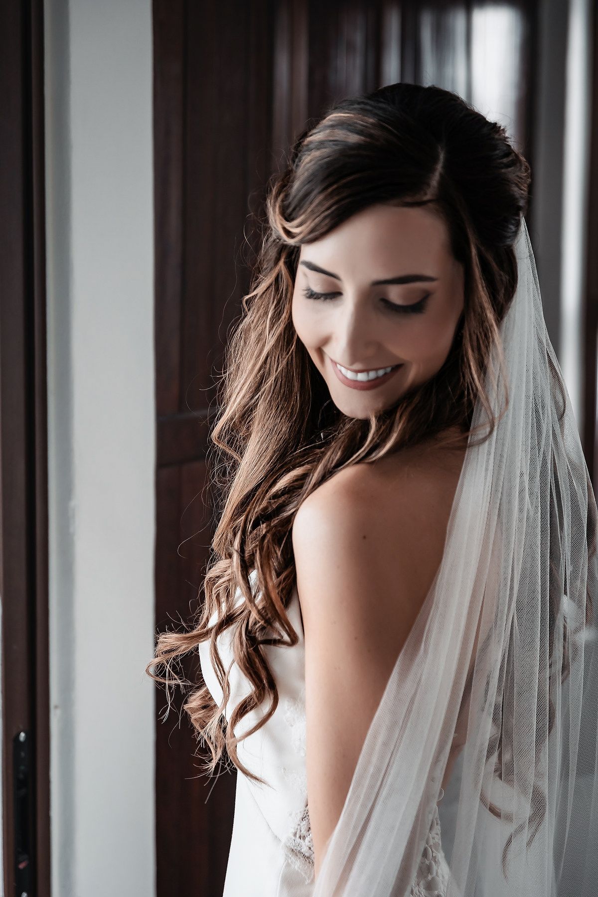 Smiling bride with long wavy brown hair and white veil looking down, wearing a strapless wedding gown near a window.