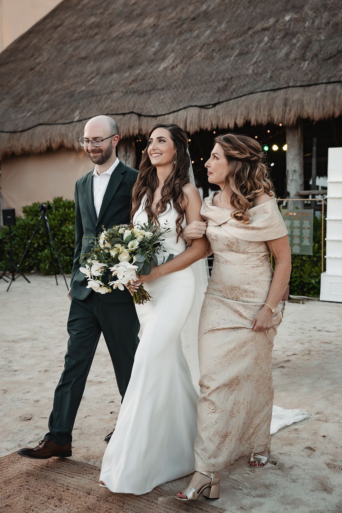 Bride in white gown holds bouquet walking with groom in dark suit and woman in gold dress on sandy beach.