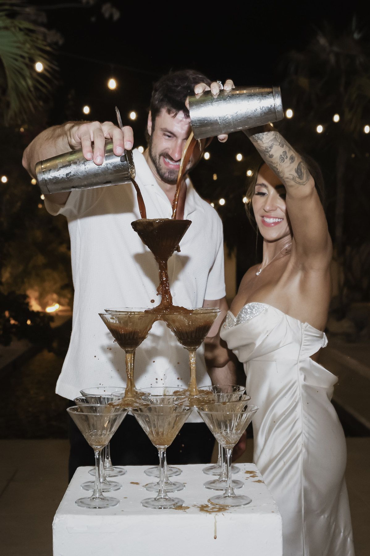 Bride and groom pouring chocolate martinis into a cocktail tower at their outdoor wedding reception at night.