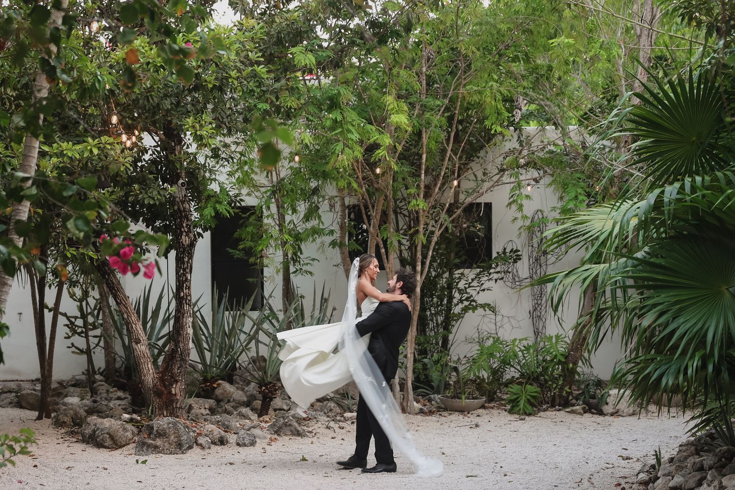 Groom lifting bride in wedding dress outdoors among lush tropical trees and palms near white building.