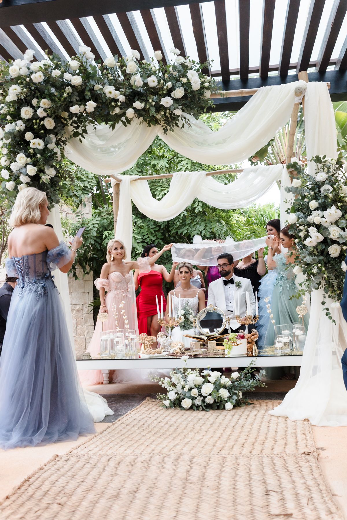 Elegant outdoor wedding ceremony under floral arch with white draping, bridesmaids in colorful gowns, and decorated altar table.
