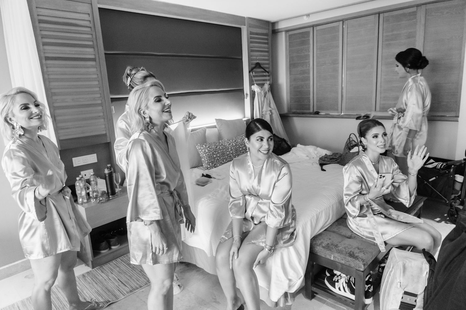 Bridesmaids in satin robes getting ready together in a hotel room, laughing and relaxing before the wedding ceremony.