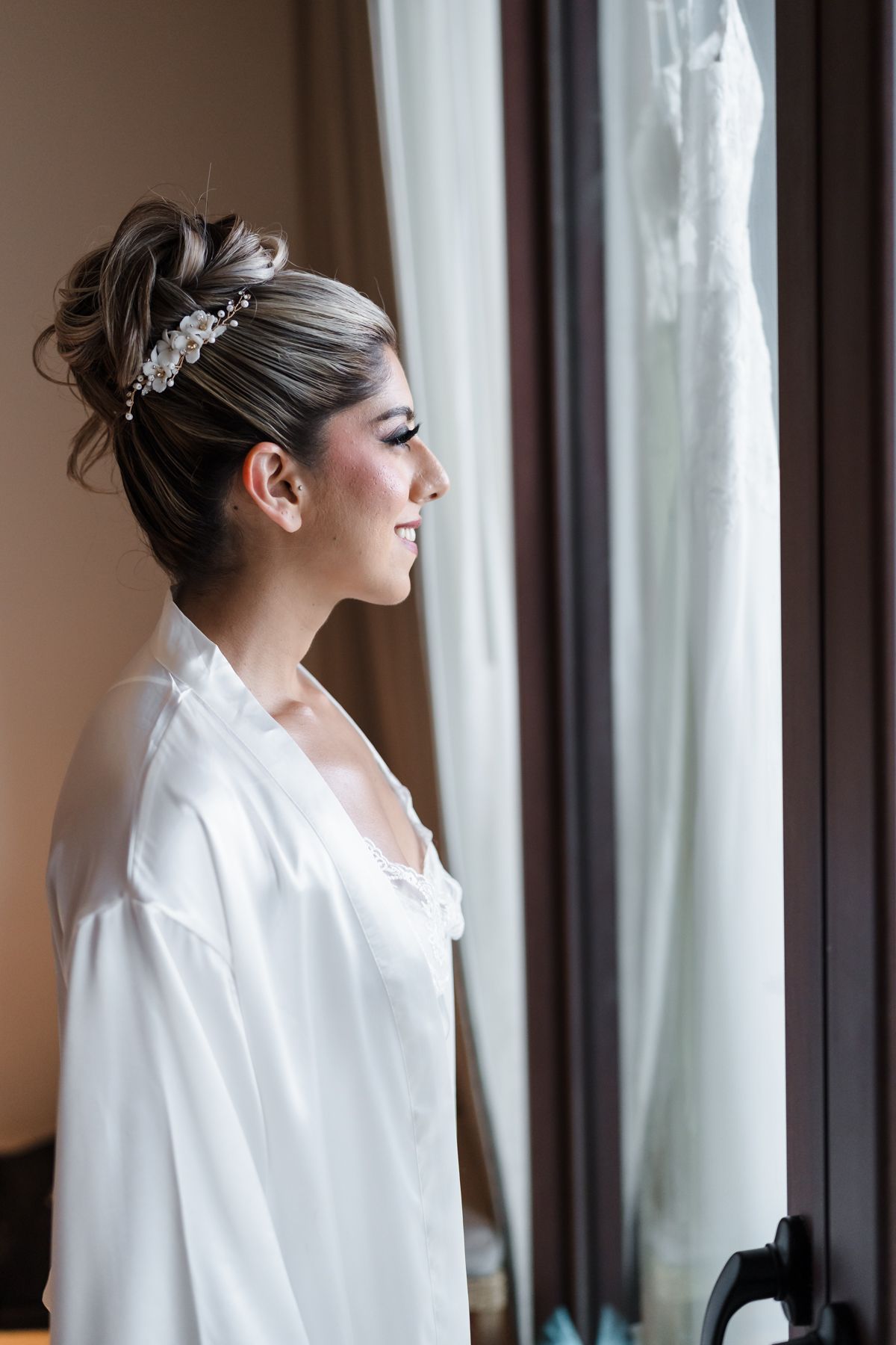 Elegant bride in white satin robe with updo hairstyle and pearl hair accessory smiling by window on wedding day.