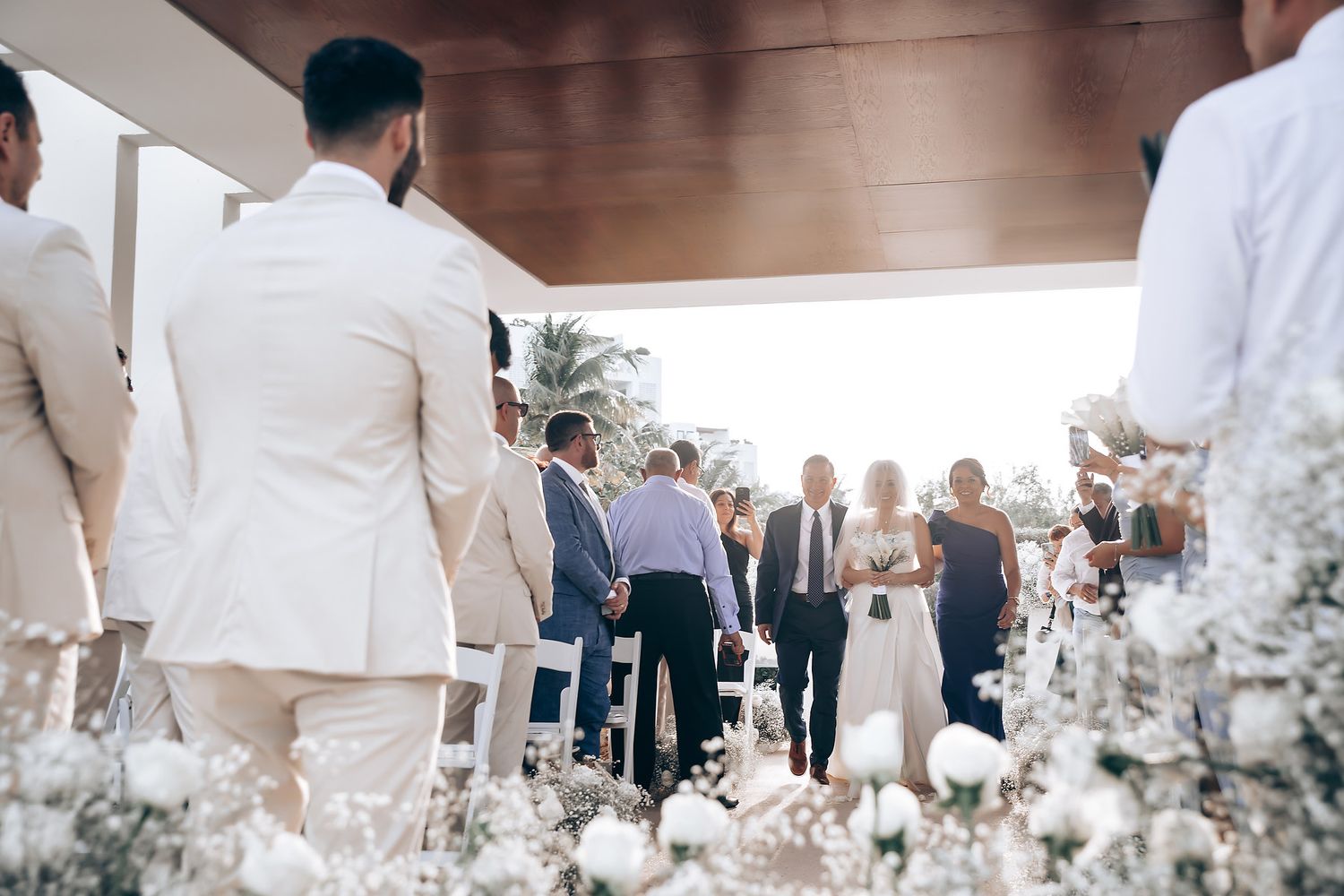 Wedding guests in white attire gather at an elegant indoor reception venue with warm lighting and decor.