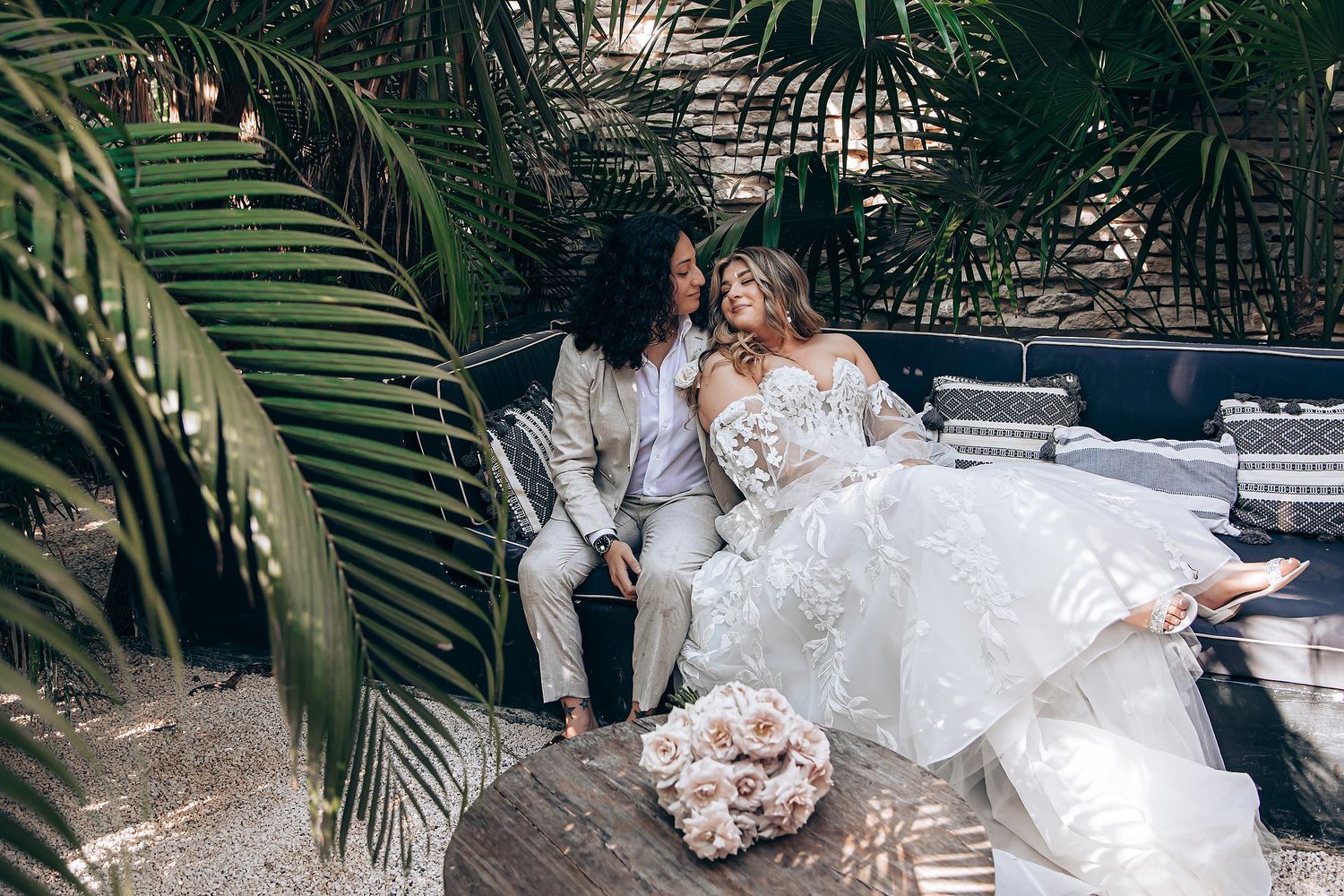 A bride in a flowing white wedding gown poses gracefully among tropical palm trees in an outdoor garden setting.