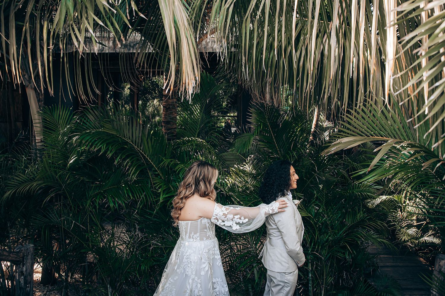 A couple in wedding attire walks through a lush tropical garden with palm fronds creating a natural archway.