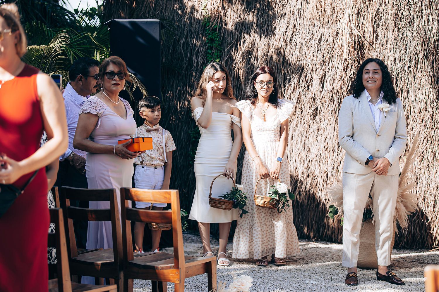 Guests stand in line at an outdoor wedding ceremony holding papers under a willow tree.