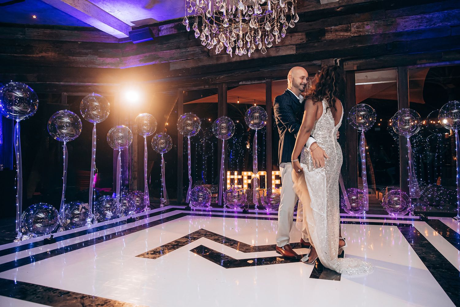 A couple shares their first dance on an elegant white dance floor under a crystal chandelier at their wedding reception.