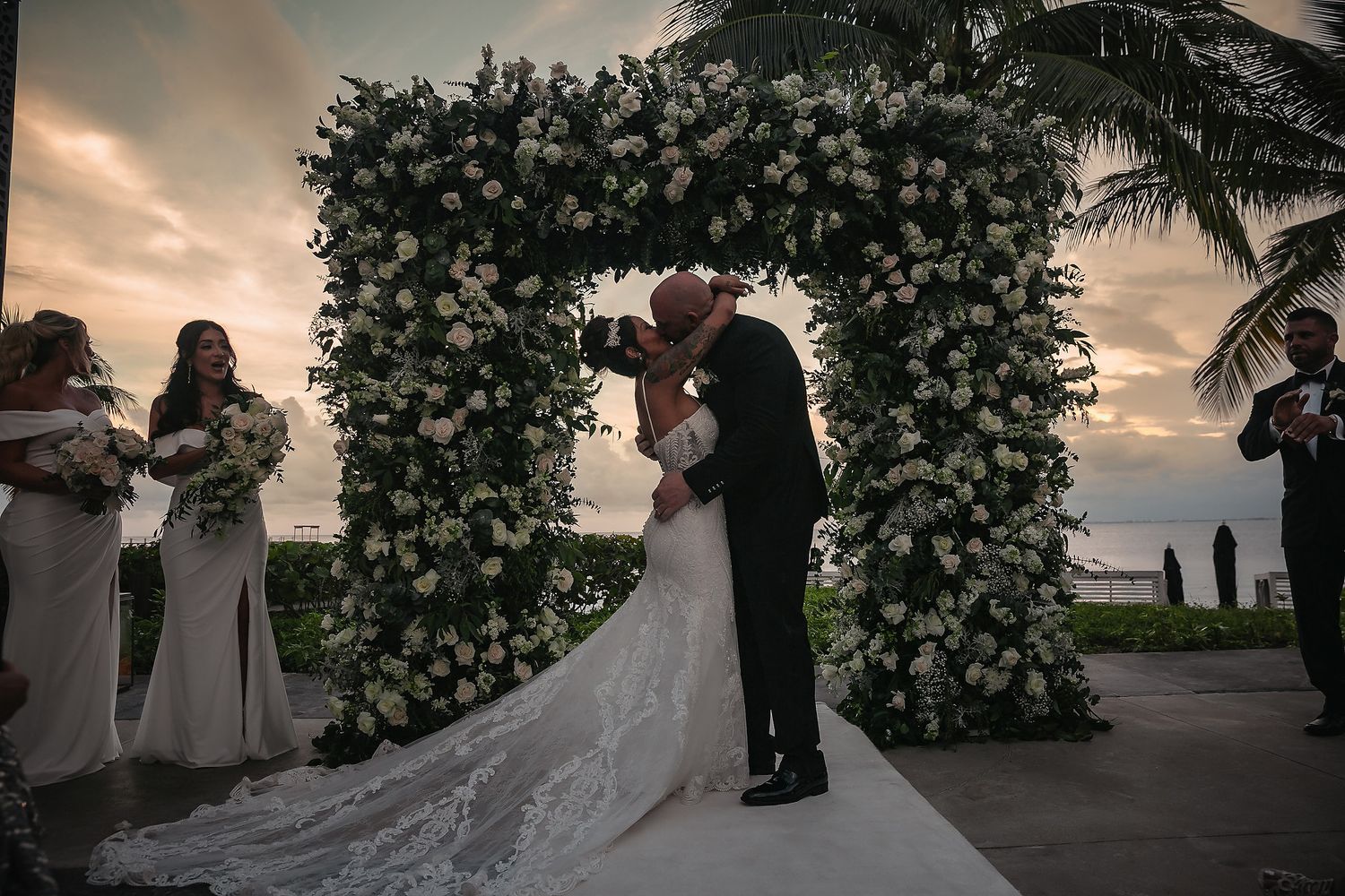 A romantic wedding couple poses at sunset under a lush floral arch during their outdoor ceremony.