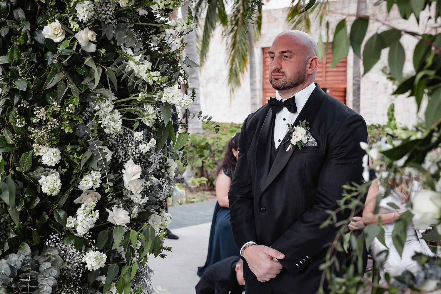 A wedding couple stands under a lush floral arch with white blooms and greenery during their outdoor ceremony.