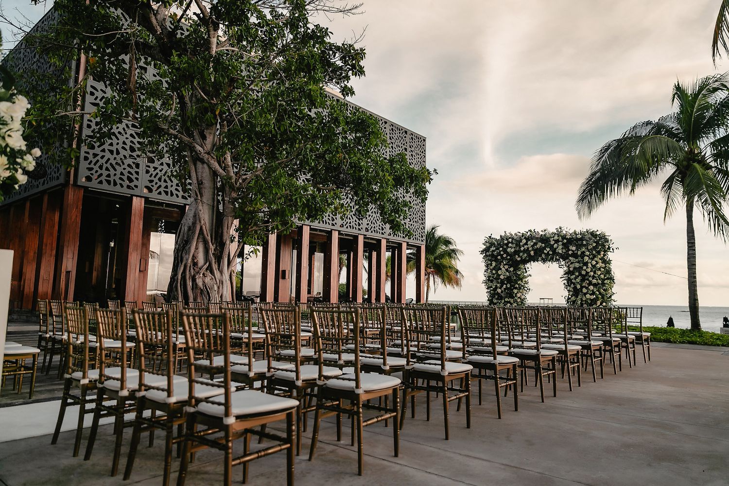 Wedding ceremony venue with white chairs arranged in rows leading to a white altar under palm trees.