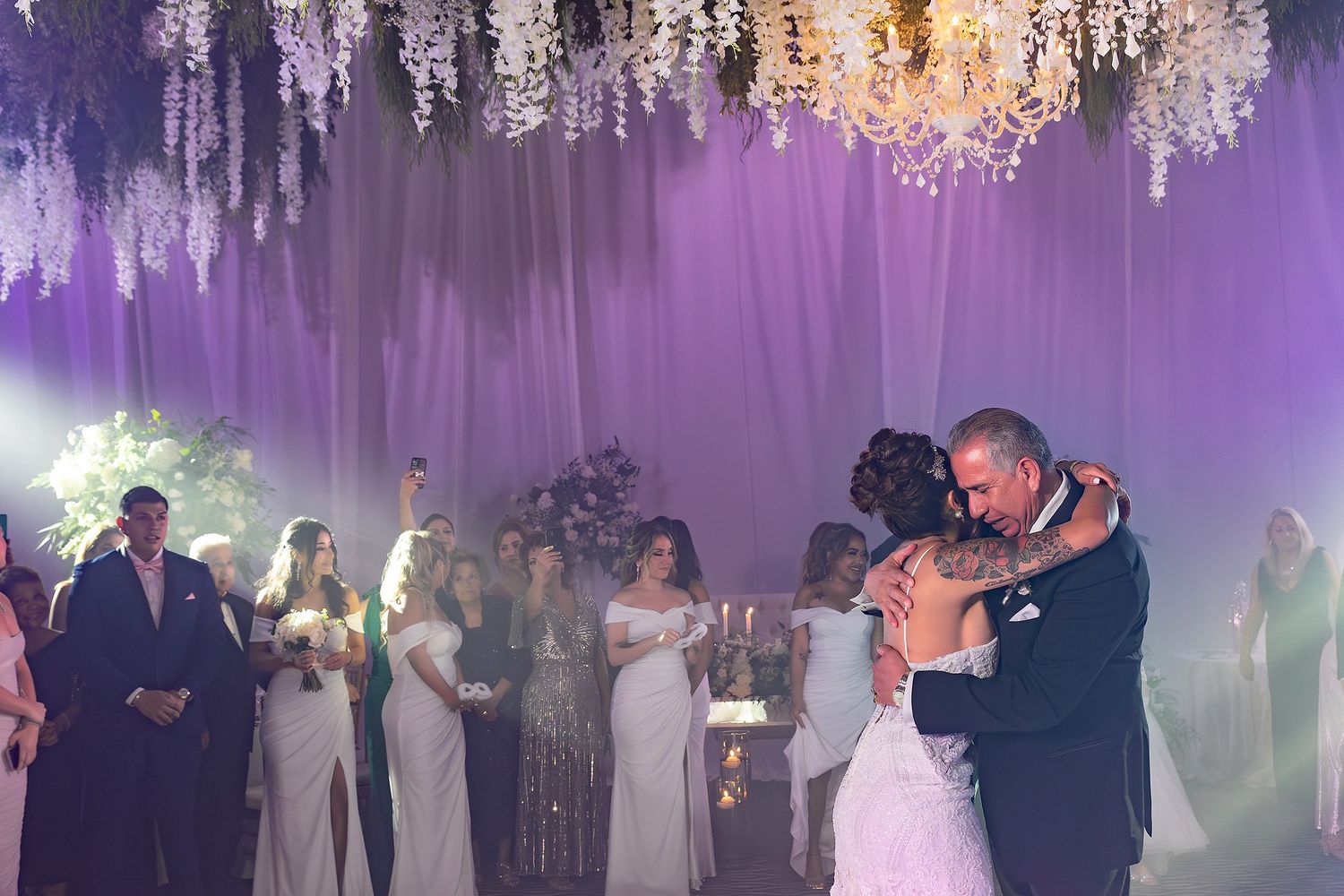 Wedding guests dance under purple lighting and hanging crystal chandeliers at an elegant reception venue.