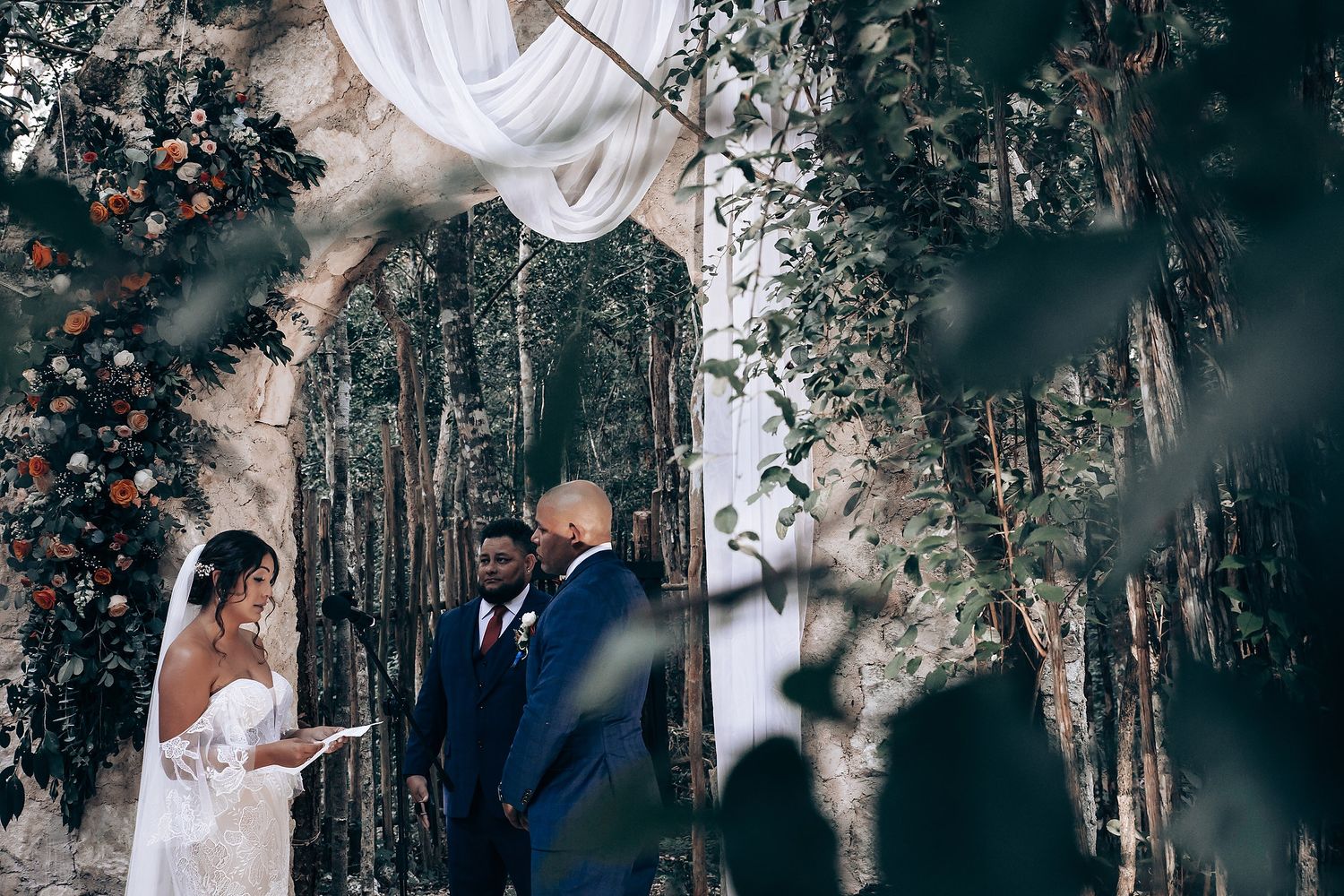 A couple exchanges vows during an intimate forest wedding ceremony beneath draped white fabric and tall trees.