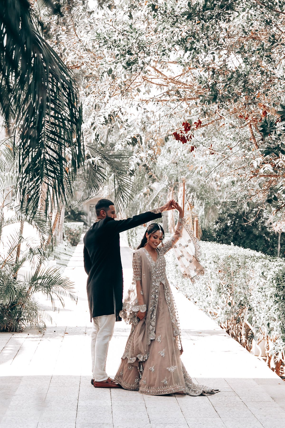 Joyful couple posing with outstretched arms on a white stone pathway surrounded by palms.
