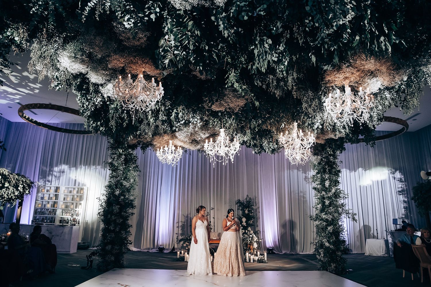 Dreamy wedding venue with white drapes, crystal chandeliers and overhead foliage creating magical ambiance.