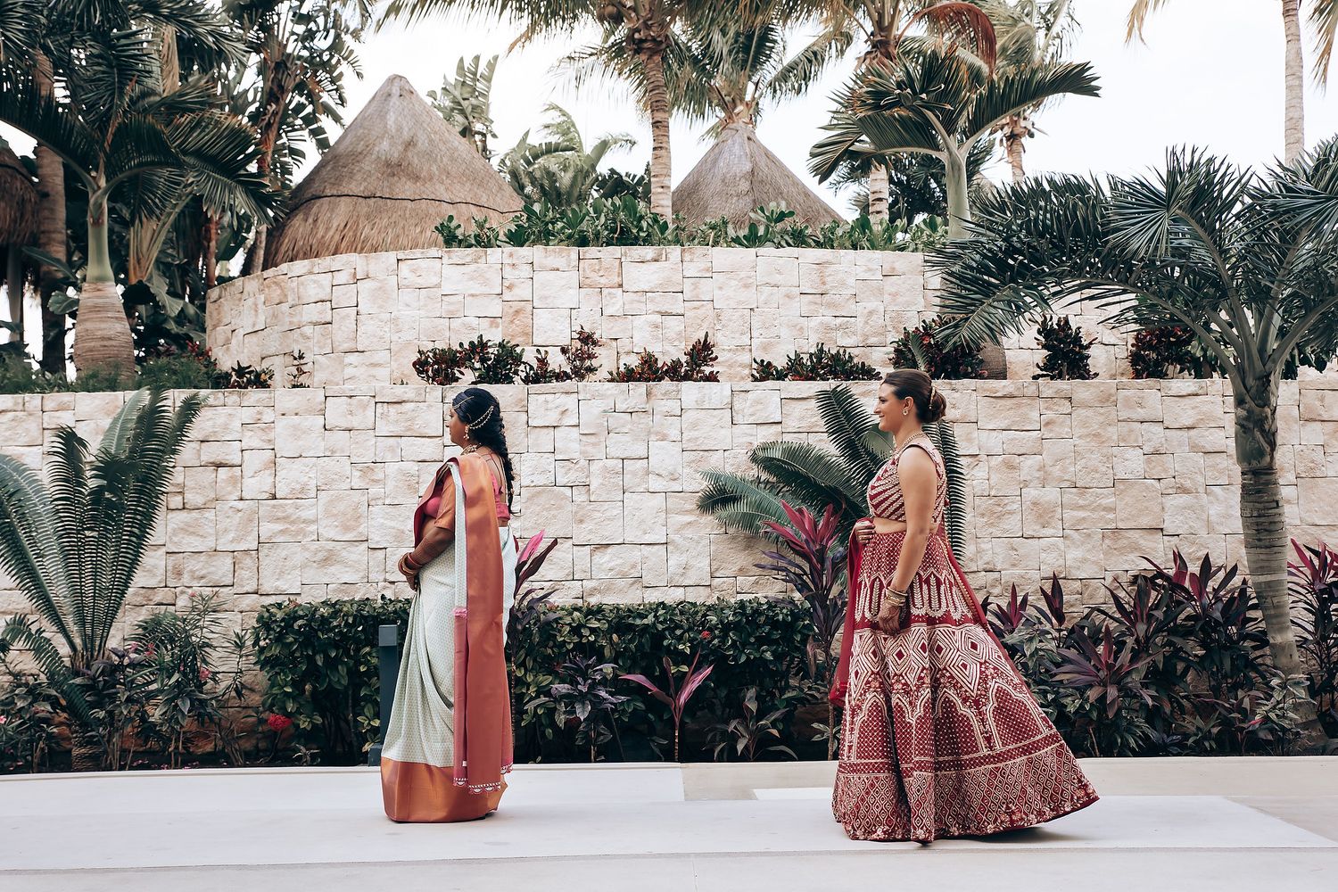 Two people in traditional Indian attire walking through a tropical garden setting with stone walls.