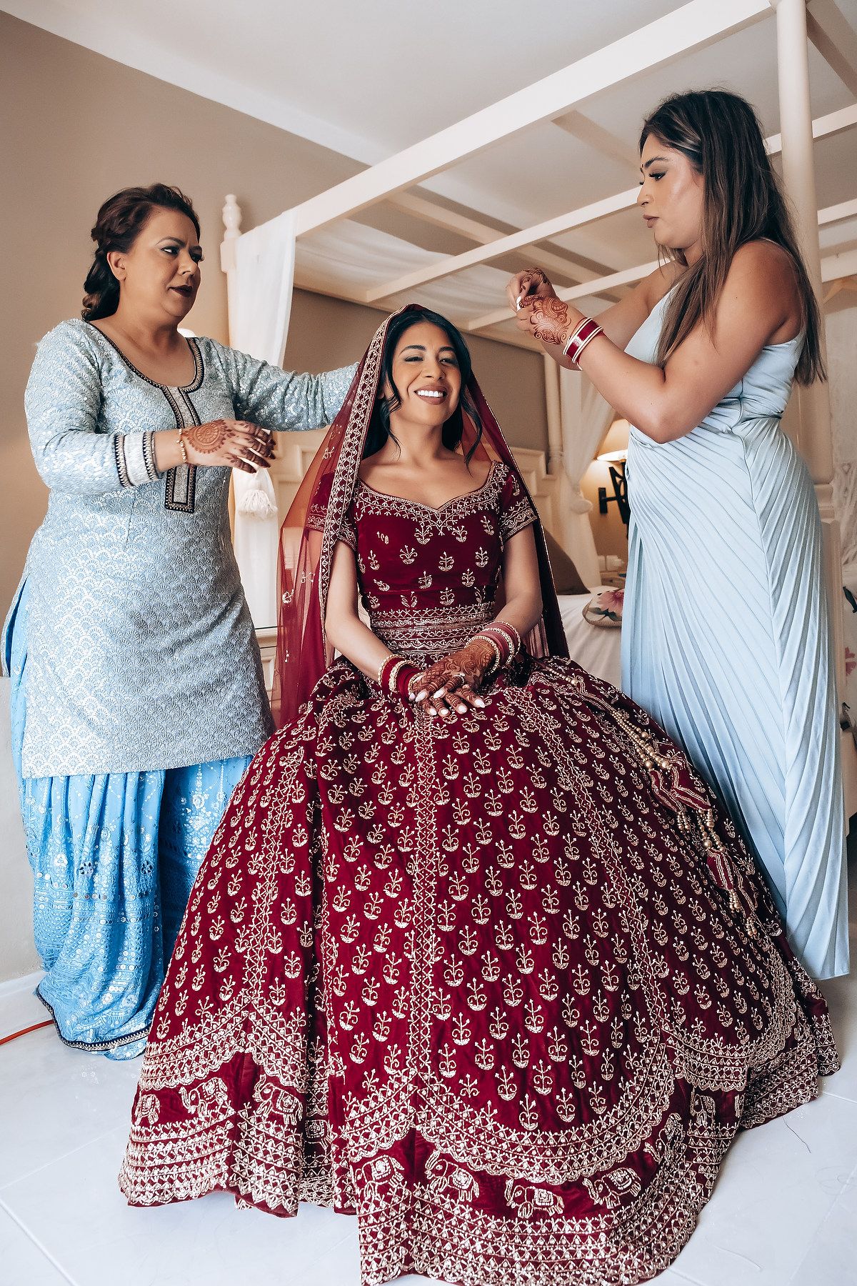 Wedding preparation scene showing styling of bride in elaborate burgundy lehenga.