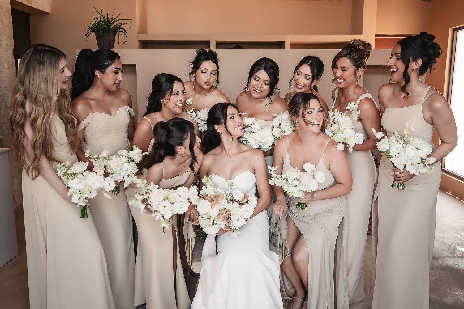 Wedding party holding white floral bouquets posed together in a light-filled venue.