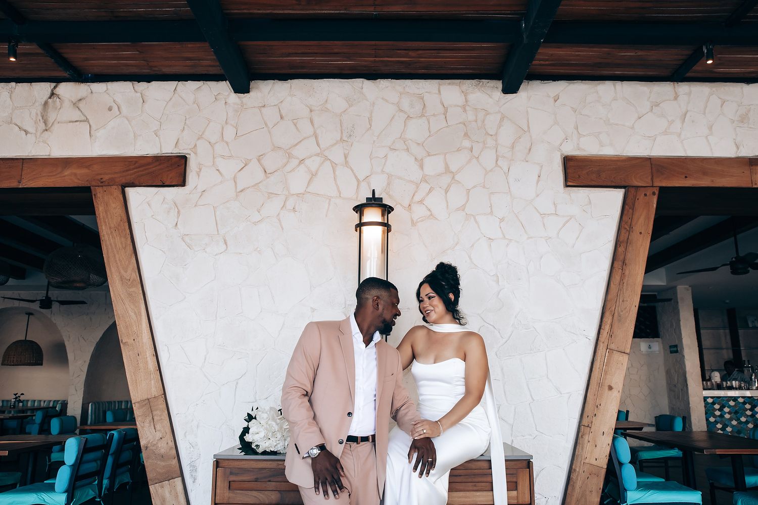 A couple poses under rustic wooden beams and wall lighting in formal attire.