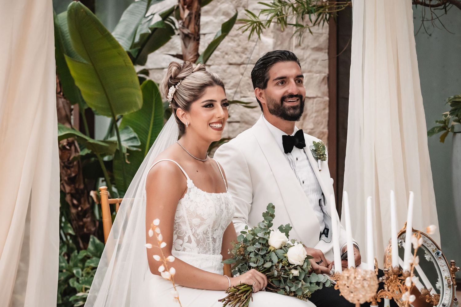 A couple shares an intimate moment during their tropical wedding ceremony with elegant decor and palm leaves.
