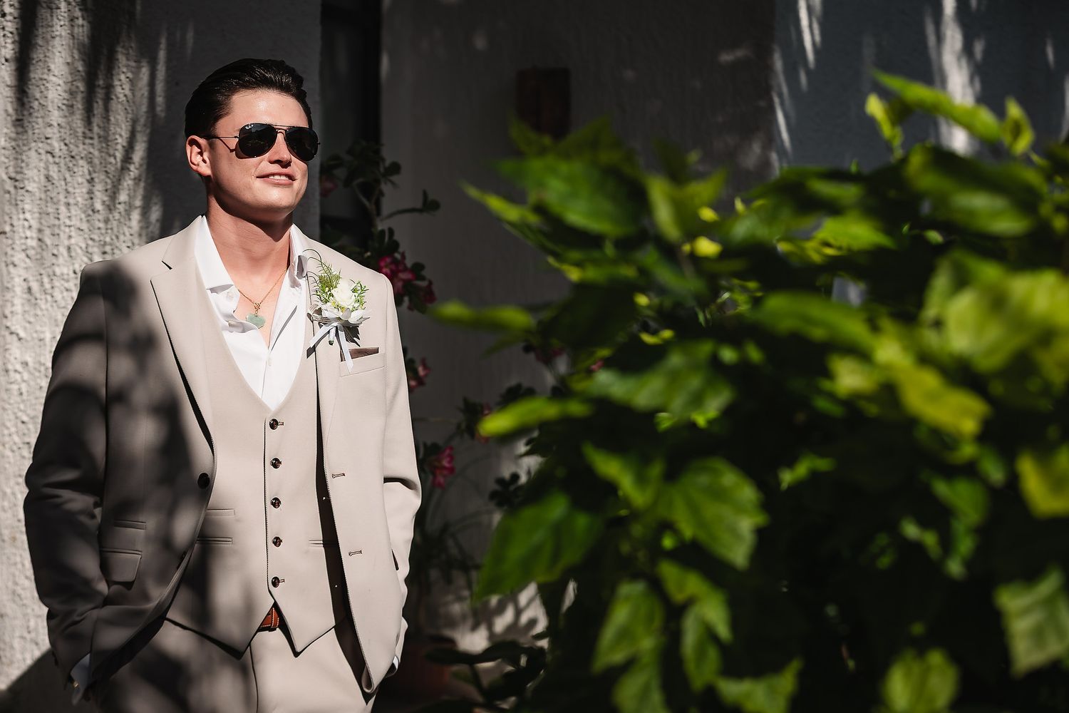 Elegant groom in light gray suit and sunglasses stands in dappled sunlight.