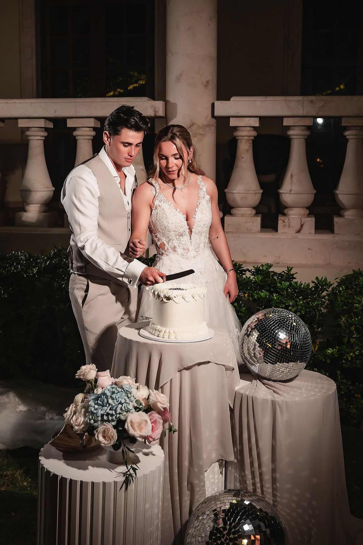 Wedding cake cutting moment captured at an elegant outdoor venue with disco balls and floral decorations.