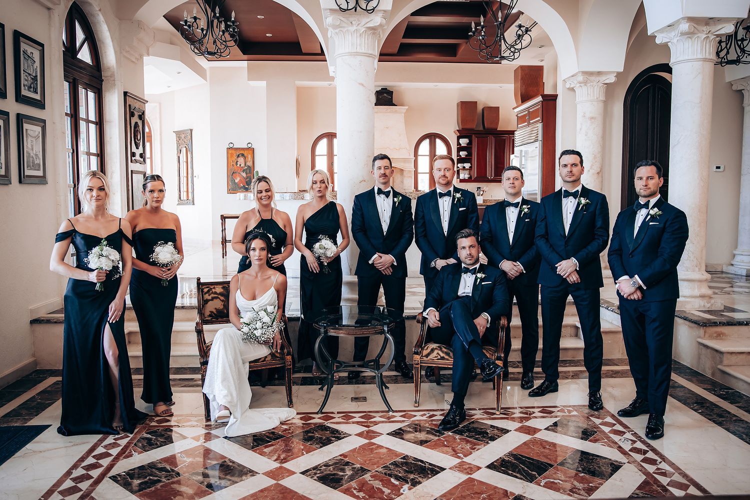Wedding party lined up in formal attire inside an elegant hallway with marble floors and arched architecture.