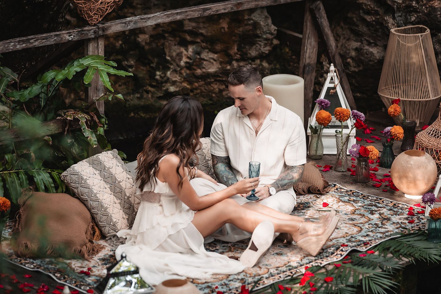 Romantic picnic scene with white decor, lanterns and rose petals on patterned blanket.
