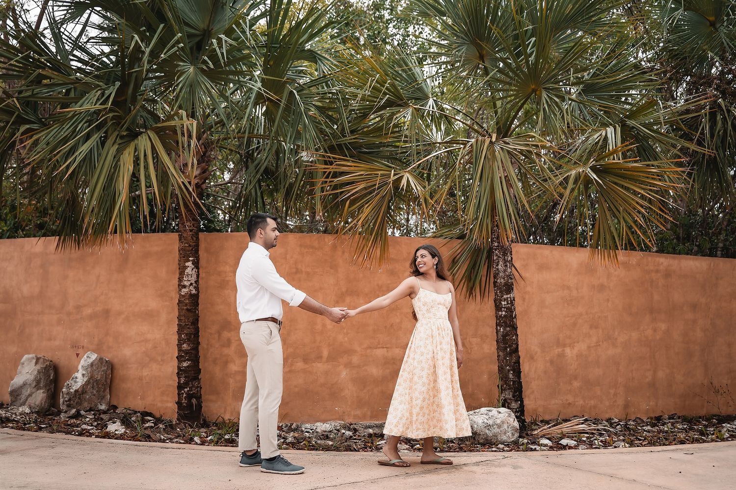 Romantic couple in white and beige outfits pose together in a tropical garden setting.