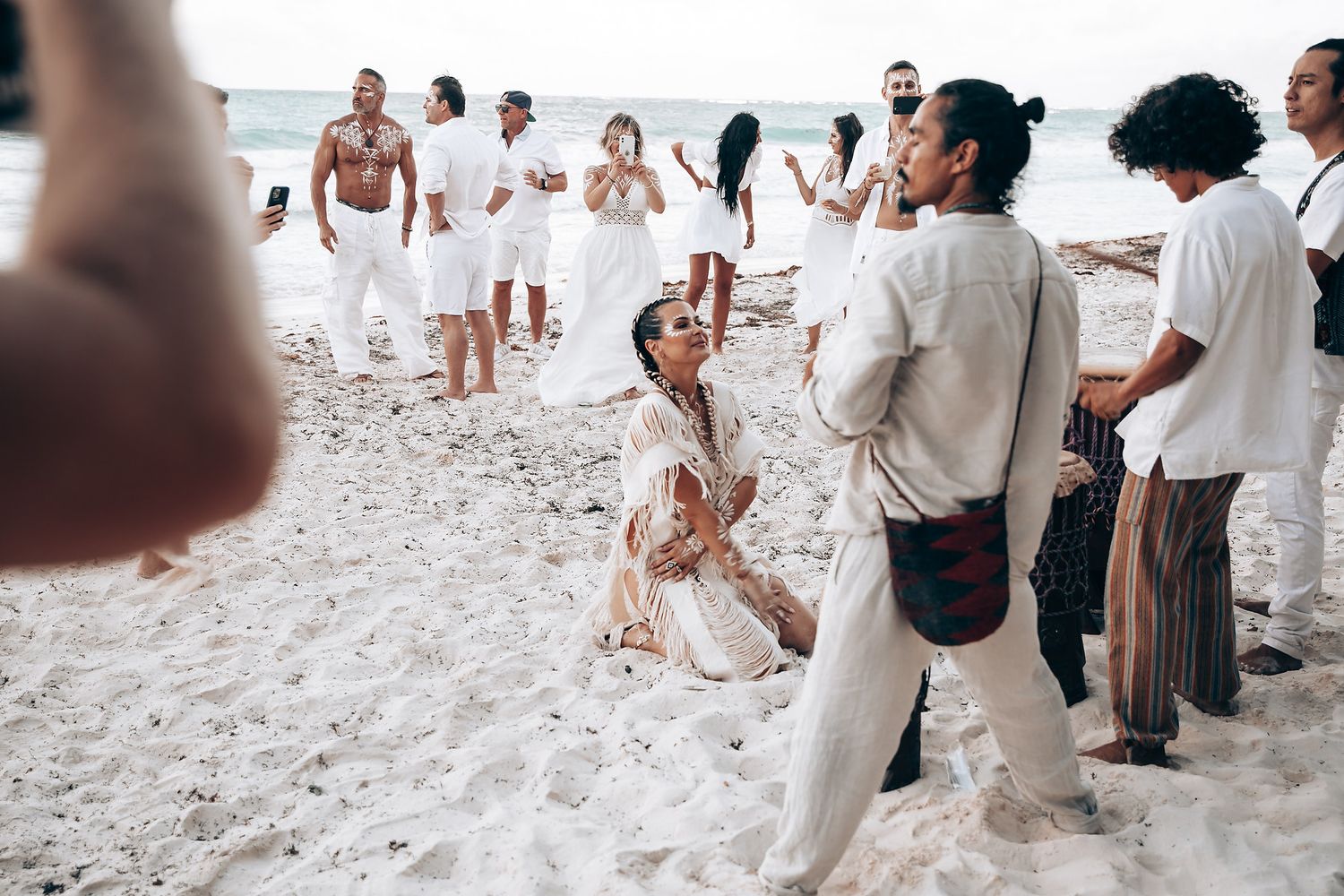 Wedding guests dressed in white gather on a sandy beach for a celebration.