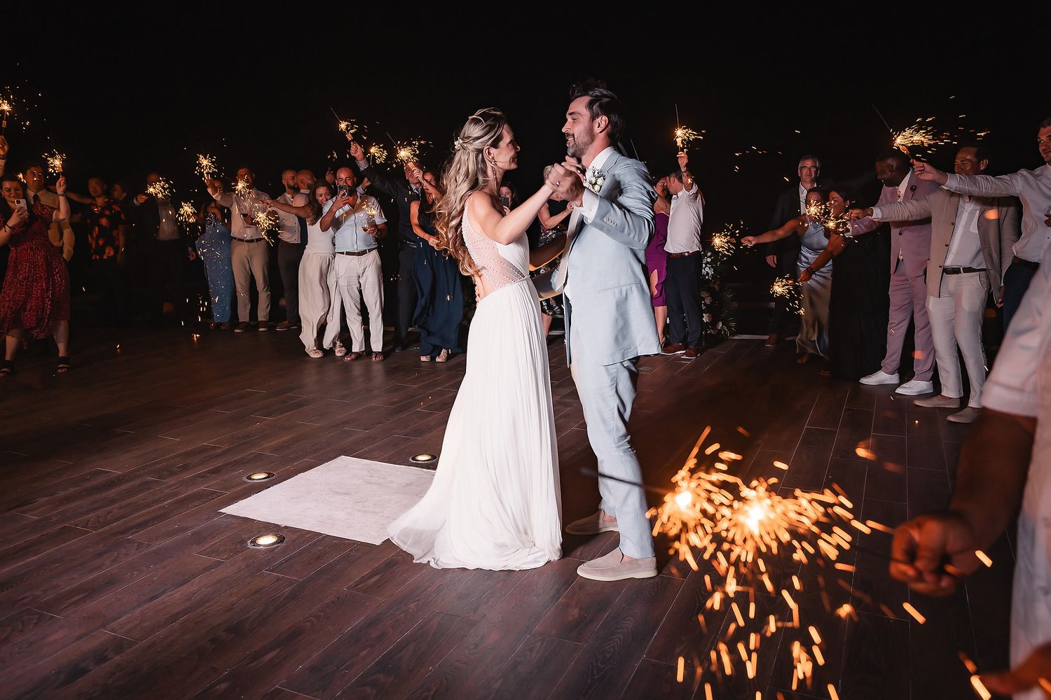 Couple shares first dance while sparklers create dramatic lighting effects on wooden deck.