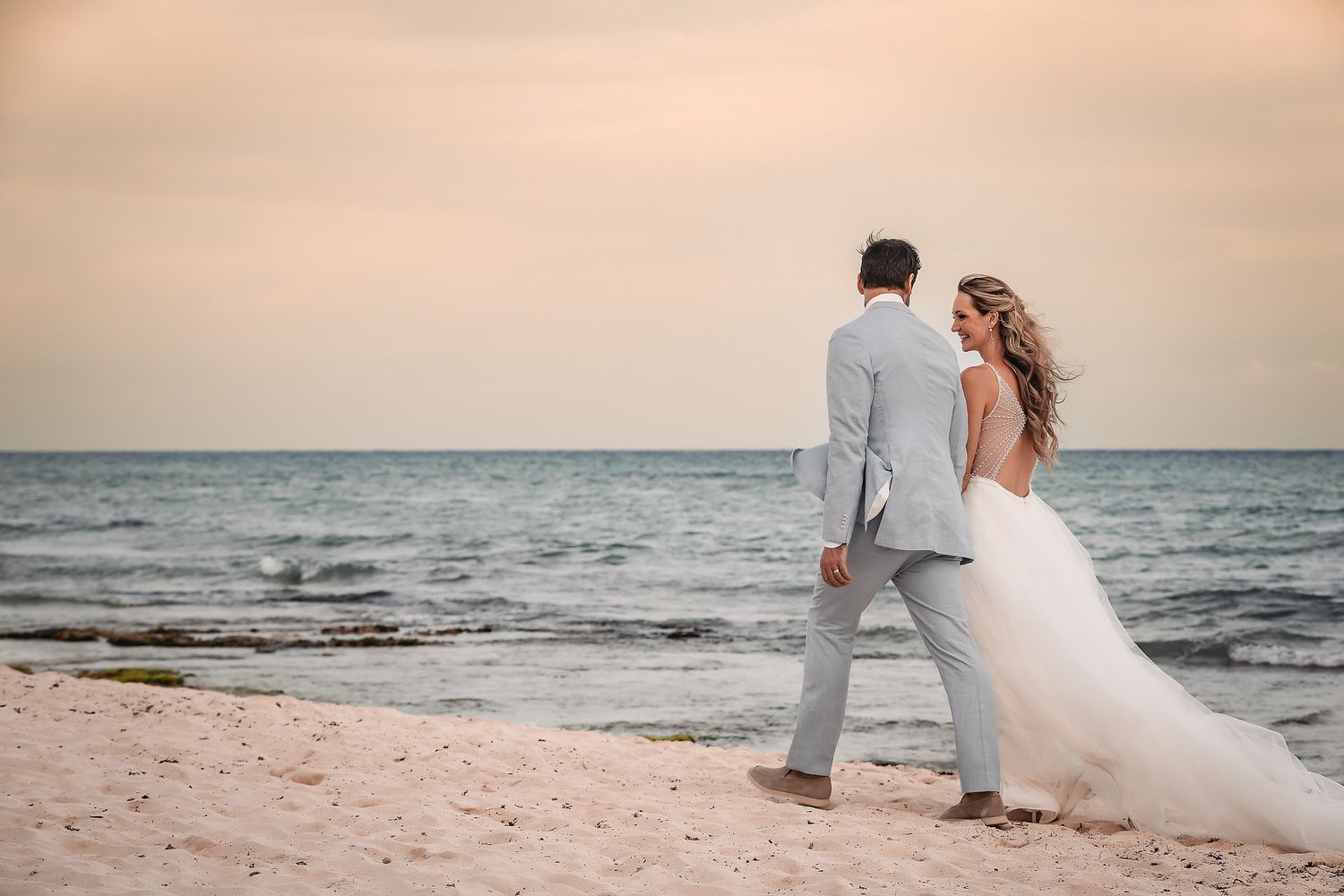 Bride in flowing white dress and groom in grey suit pose on beach during sunset.
