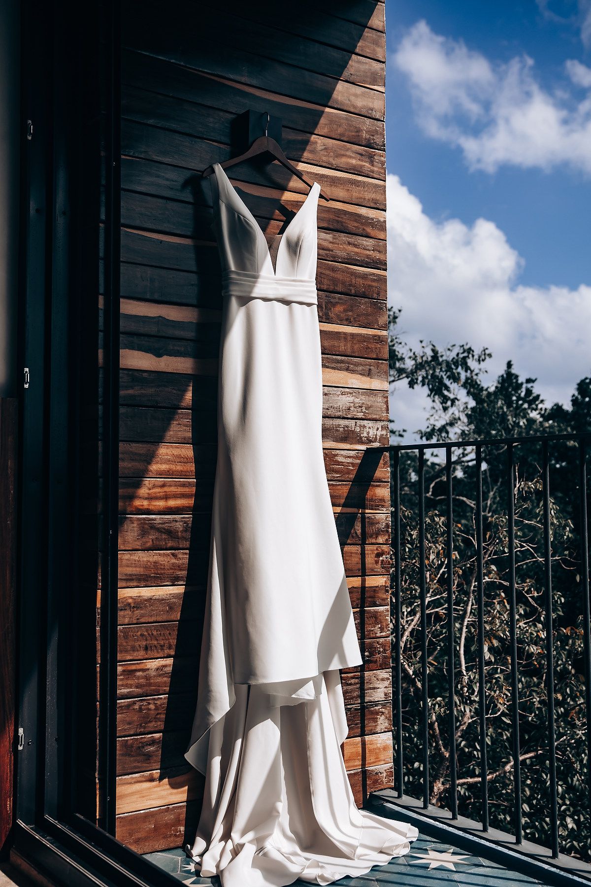 White wedding gown displayed against rustic wood paneling with blue sky and trees in background.
