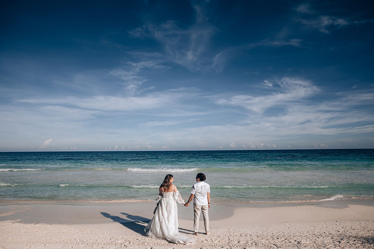 A couple stands together on a pristine beach looking out at the turquoise ocean and blue sky.