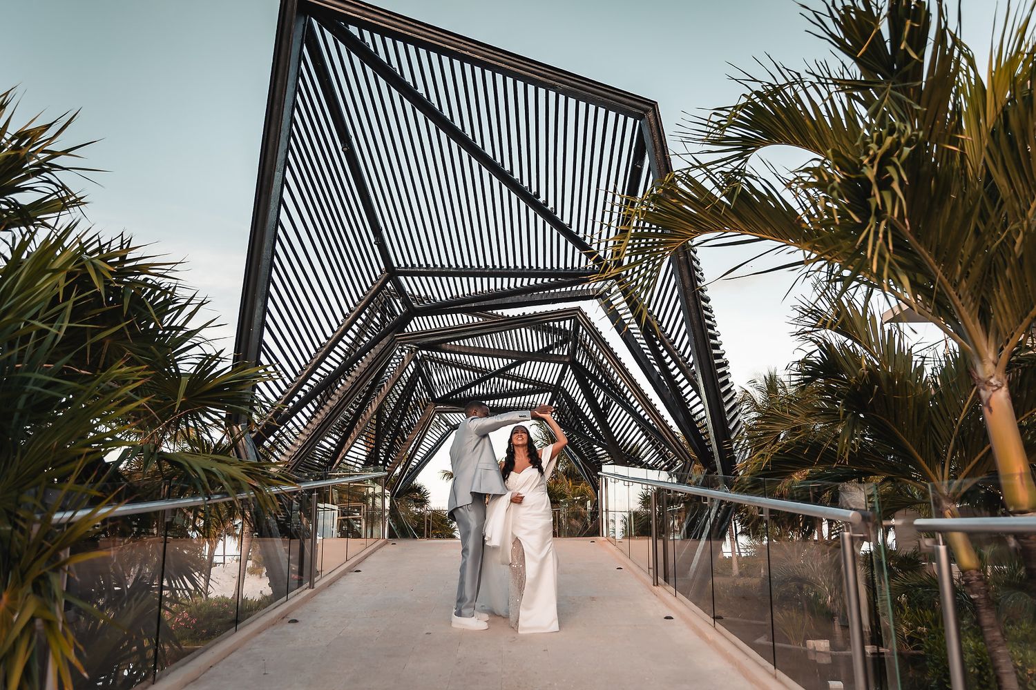 A couple walks along a palm-lined walkway with modern architectural elements at a tropical wedding venue.