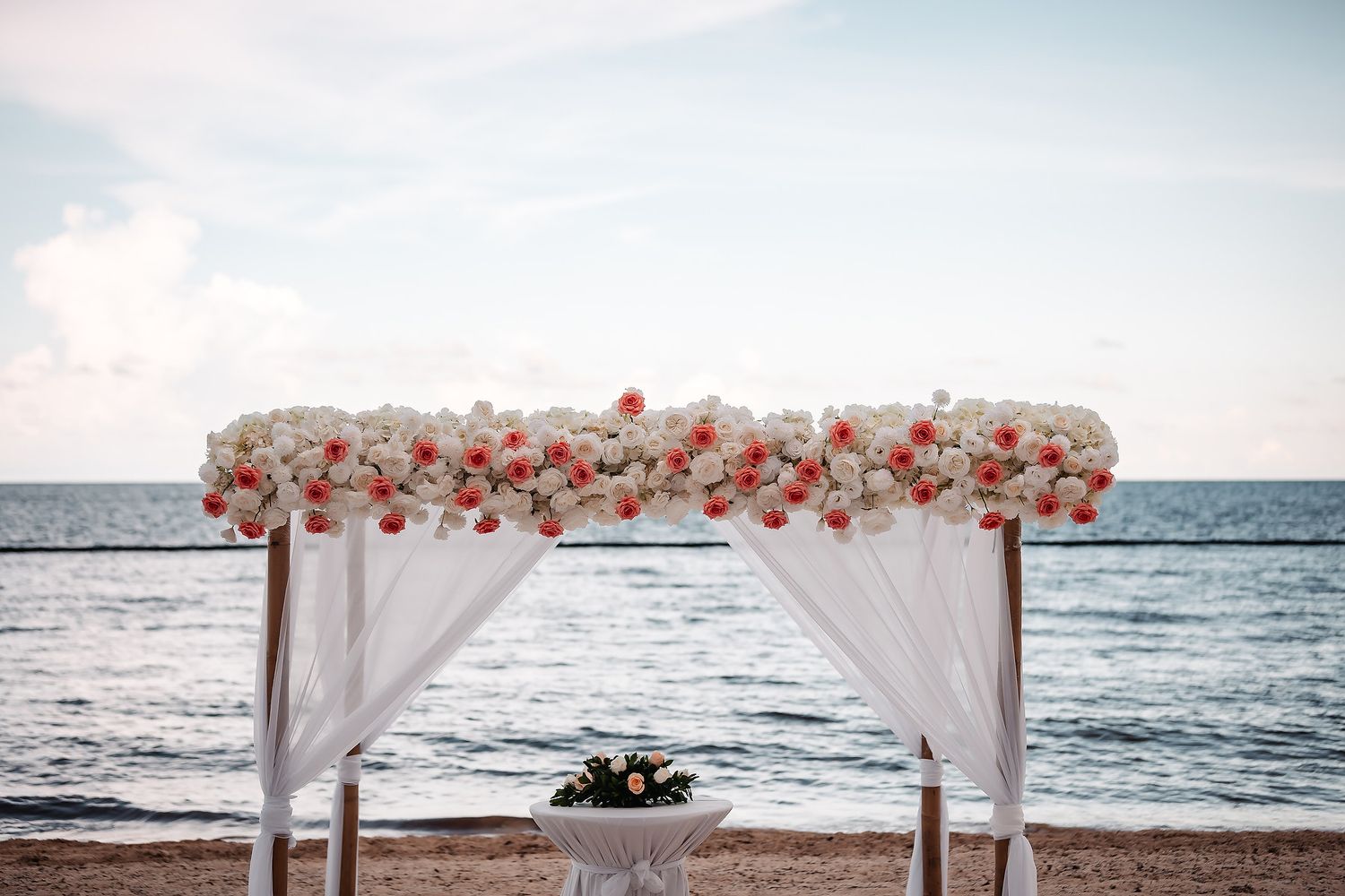 White wedding arch decorated with coral and white flowers overlooking the ocean at sunset.