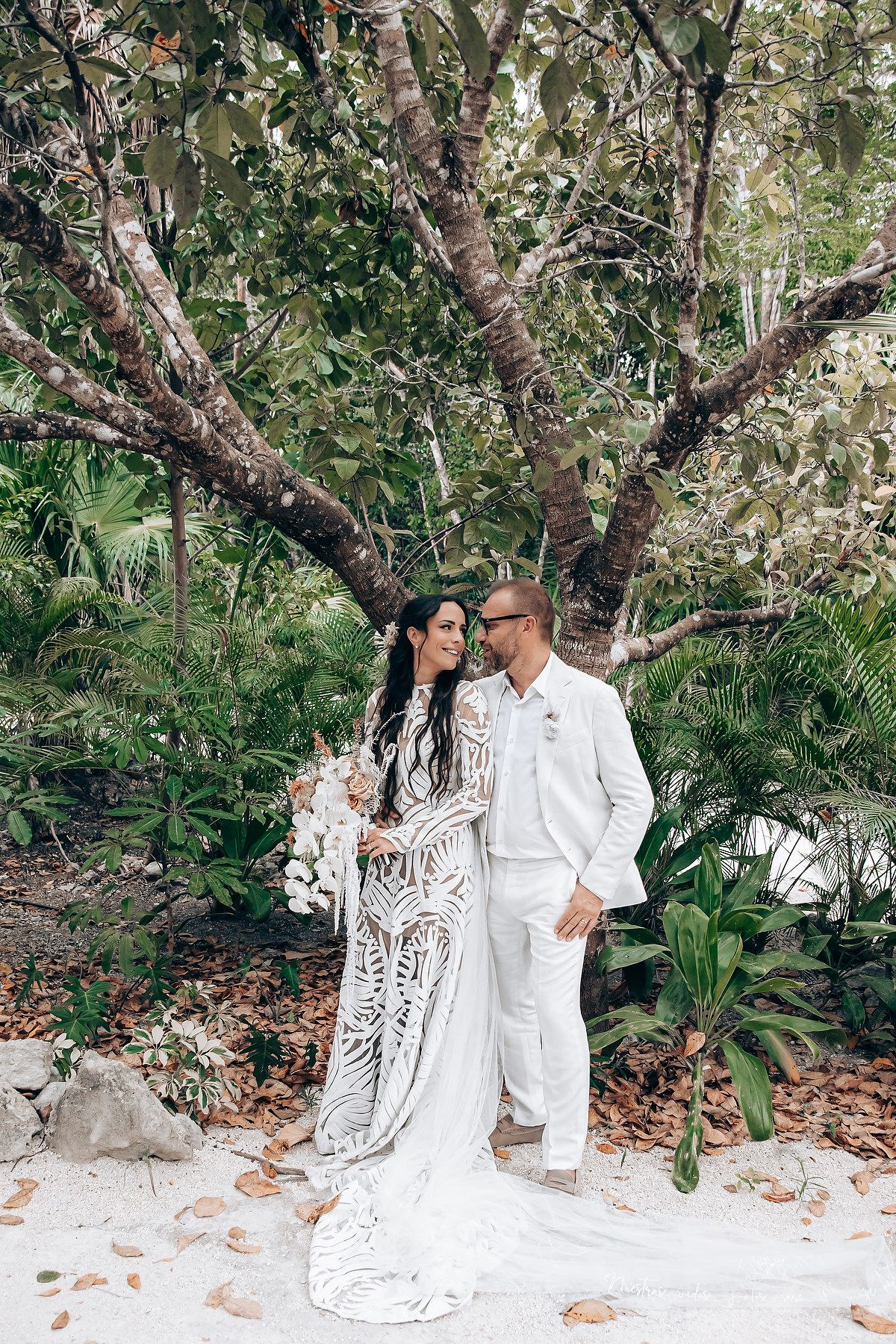 A couple in coordinated white outfits pose together in a lush garden setting with tropical foliage and trees.