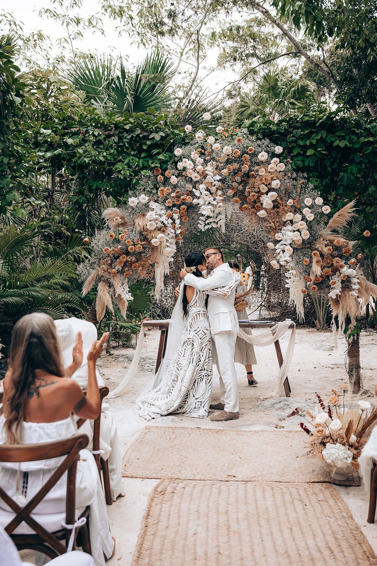 Wedding ceremony in progress under natural archways decorated with dried flowers and palm trees.