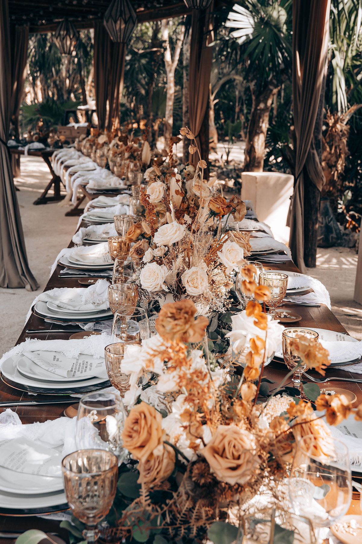 Elegant outdoor wedding reception table setting with white place settings and peach floral centerpieces at sunset.