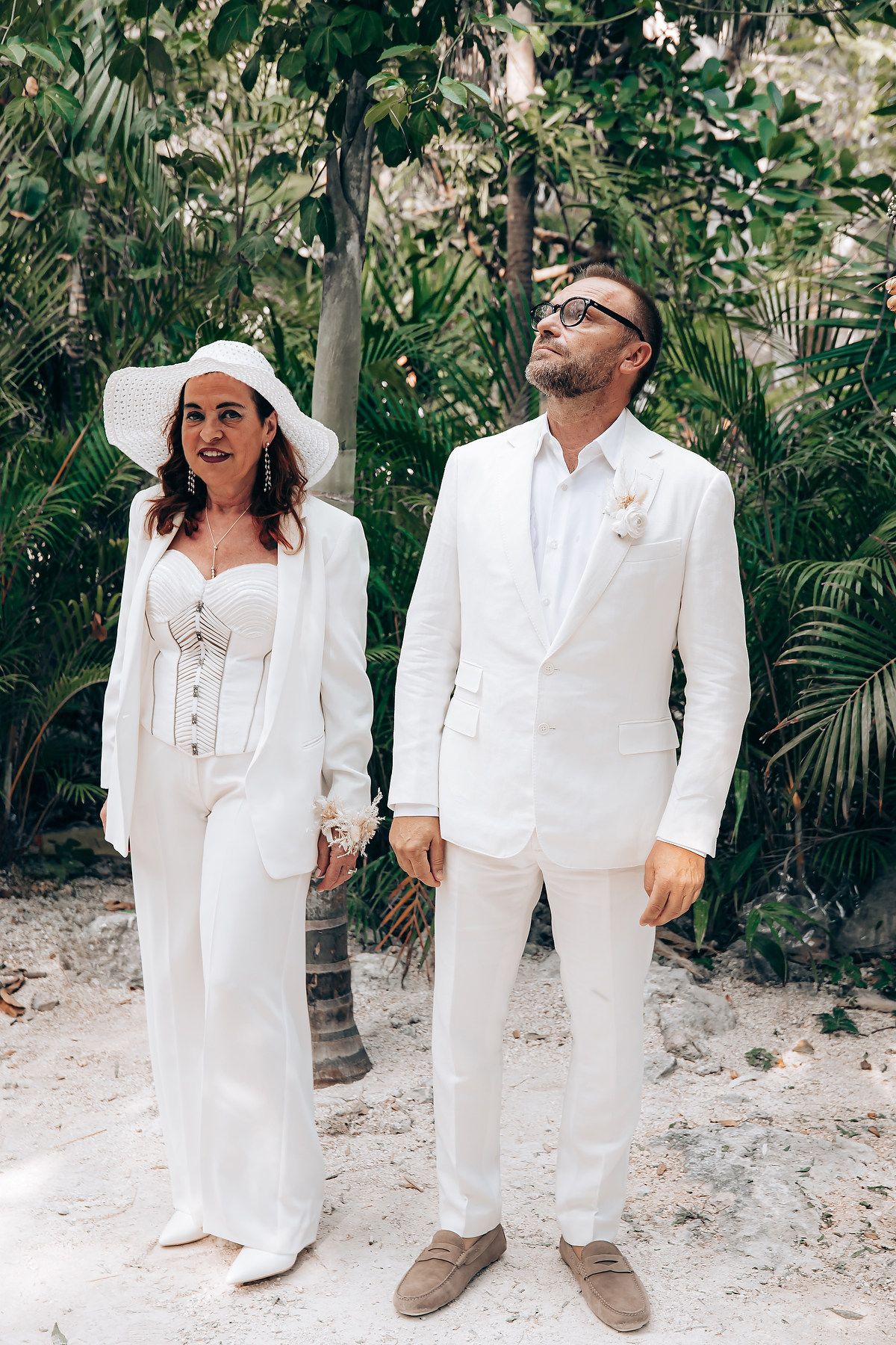 A couple in matching white outfits poses playfully together on a tropical beach path with lush greenery.