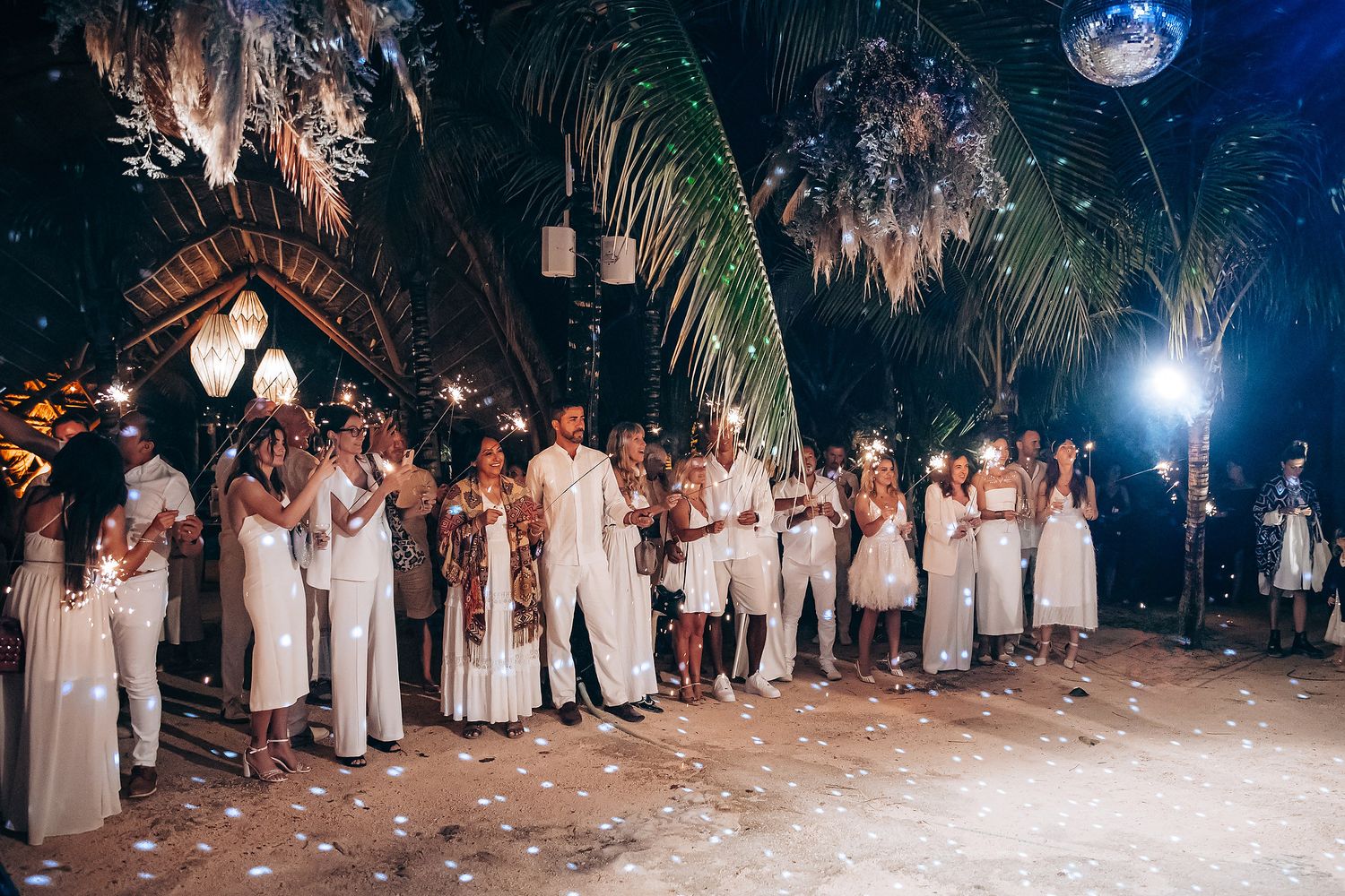 Wedding guests form a line at night under outdoor lighting and palm trees during a reception celebration.