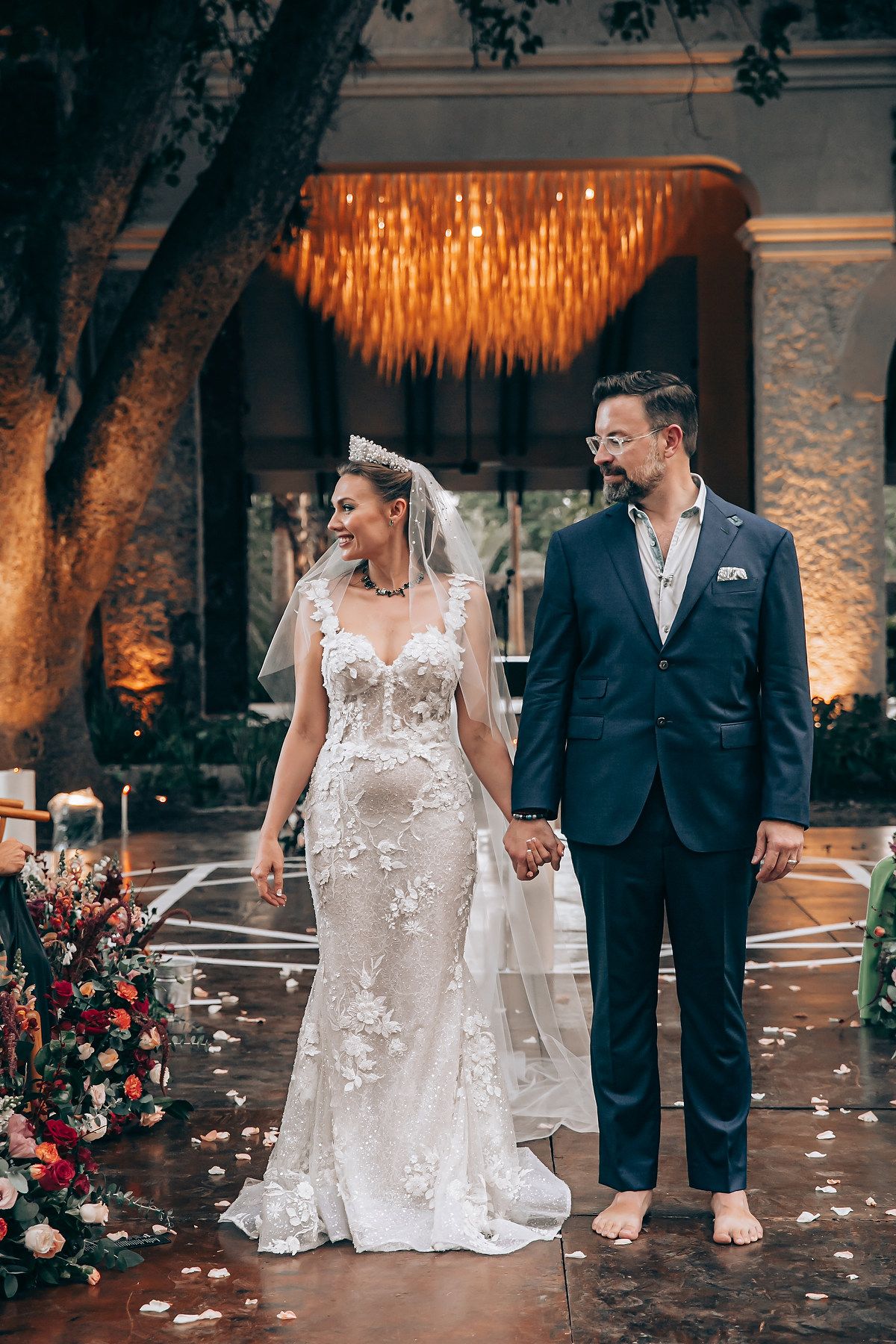 A couple in wedding attire share a joyful moment on their special day surrounded by romantic lighting and rose petals.