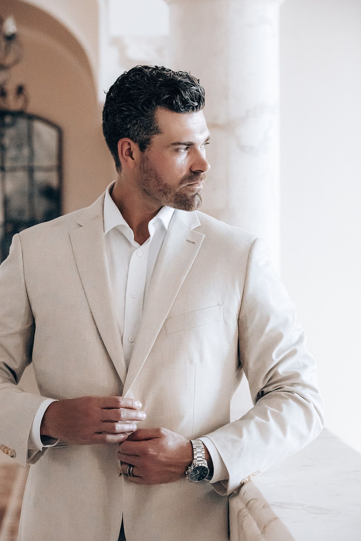 A person wearing a beige suit and white dress shirt adjusts their cuff while standing in an elegant interior space.