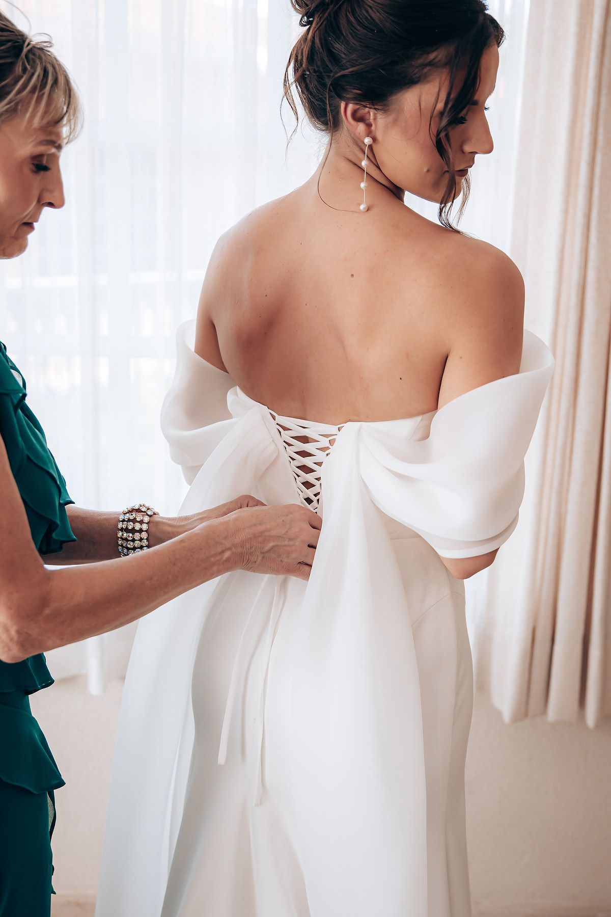 Someone helps lace up the back of an elegant white wedding dress with corset-style detailing in a bright room.