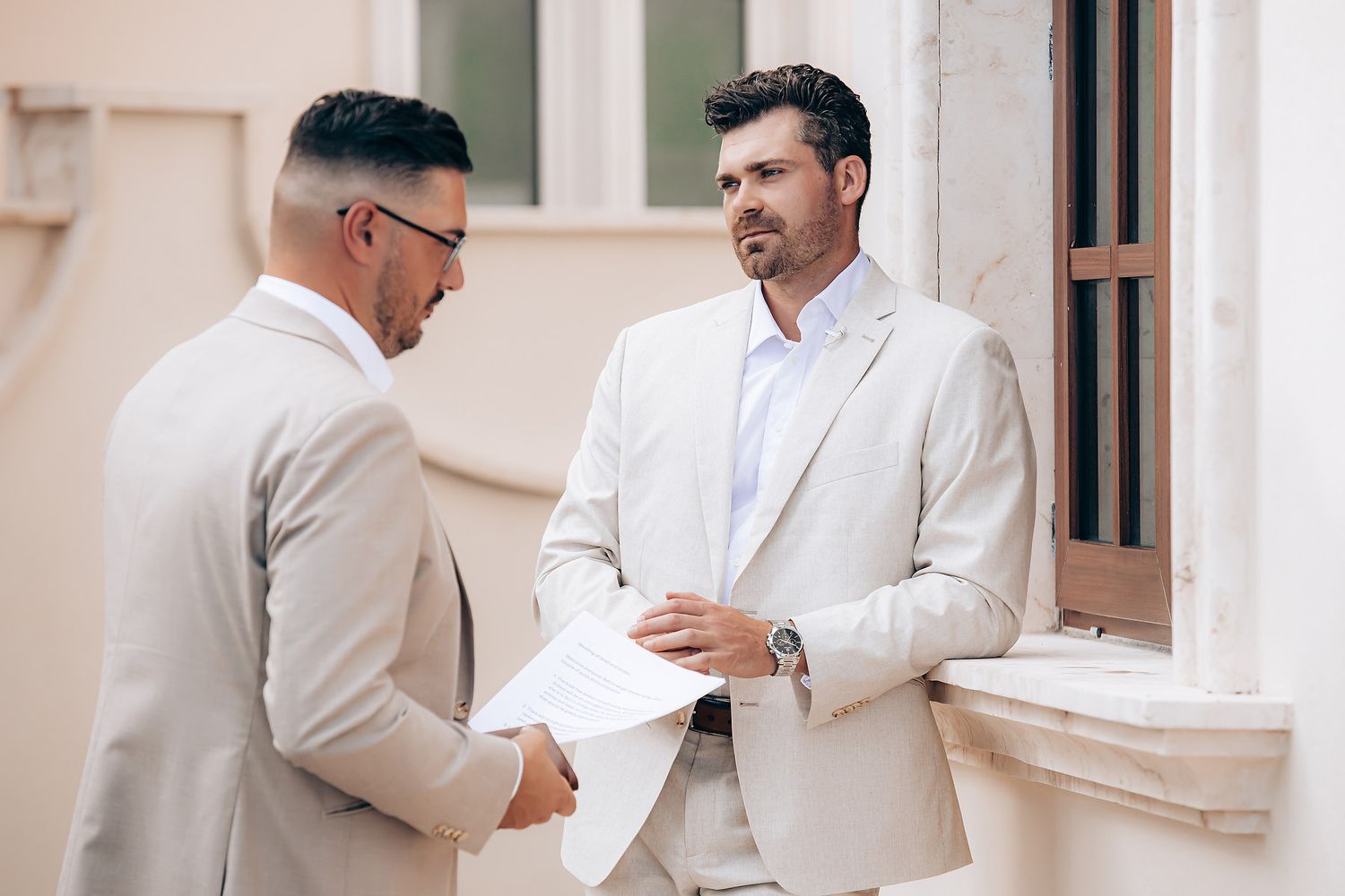 Two people in light colored suits having a conversation in an elegant white hallway.