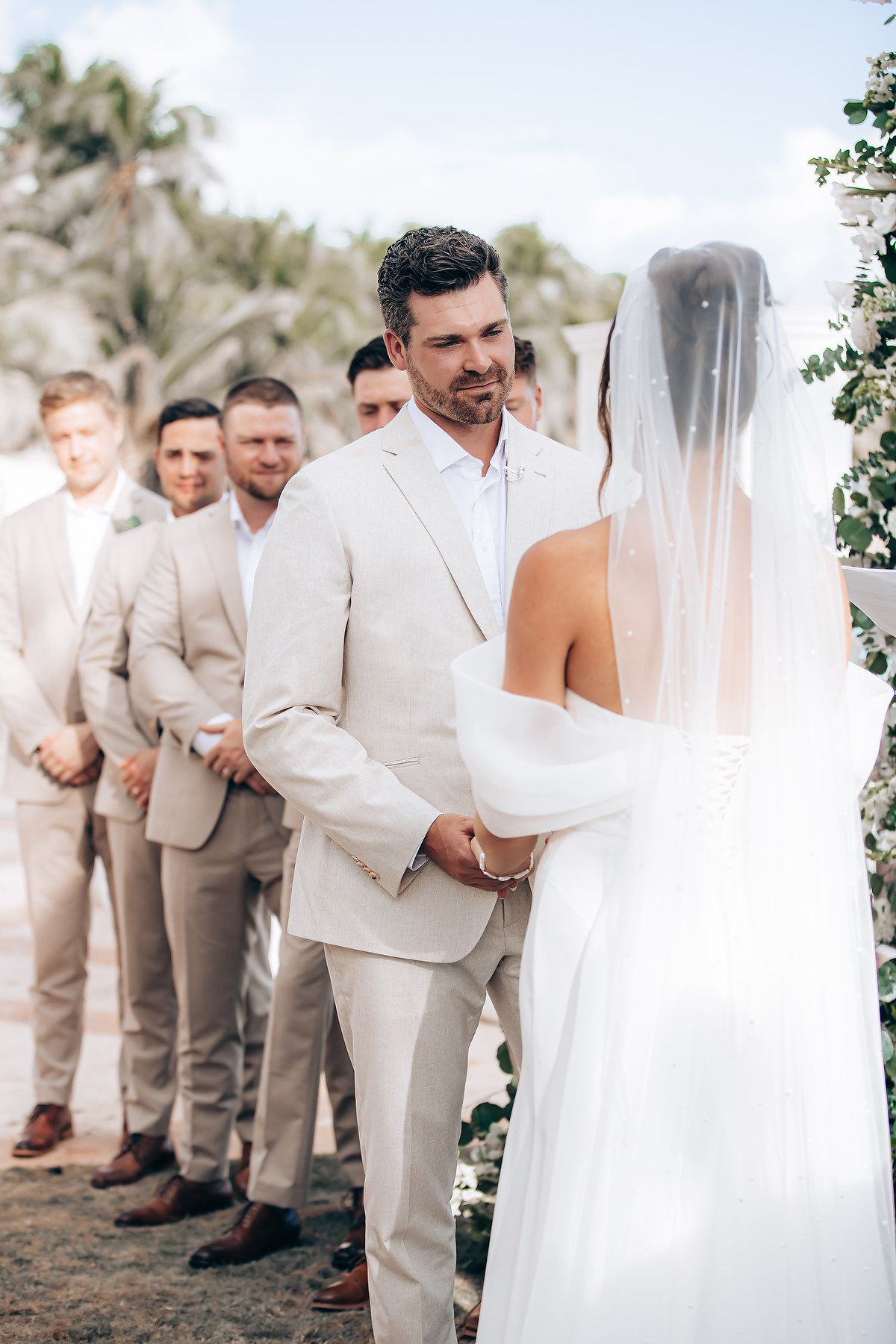 Wedding party in light beige and white attire poses together outdoors during a romantic ceremony.