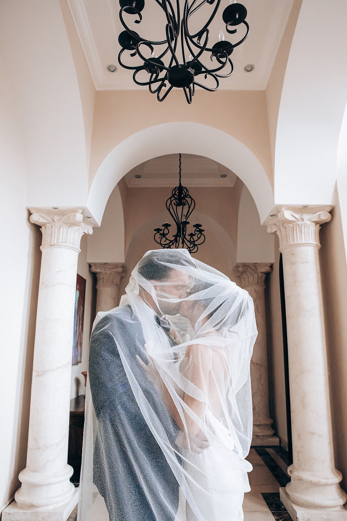 A couple embraces under a sheer white veil in an elegant columned archway with black chandeliers overhead.