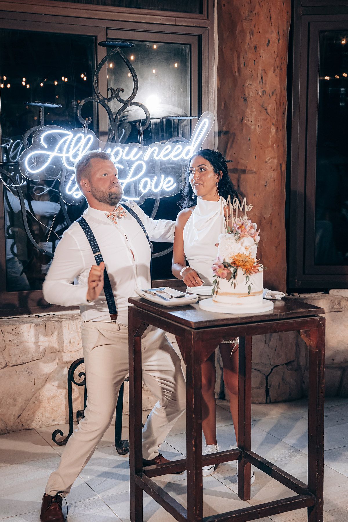 Wedding cake cutting moment under neon 'All we need is Love' sign.
