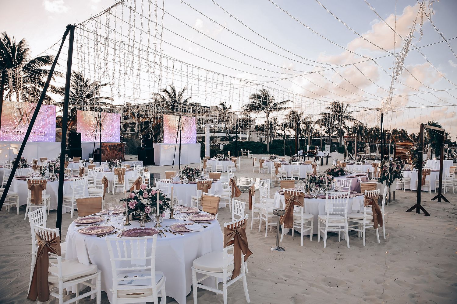 Panoramic view of a beachfront wedding venue with white tables, palm trees and hanging lights at dusk.