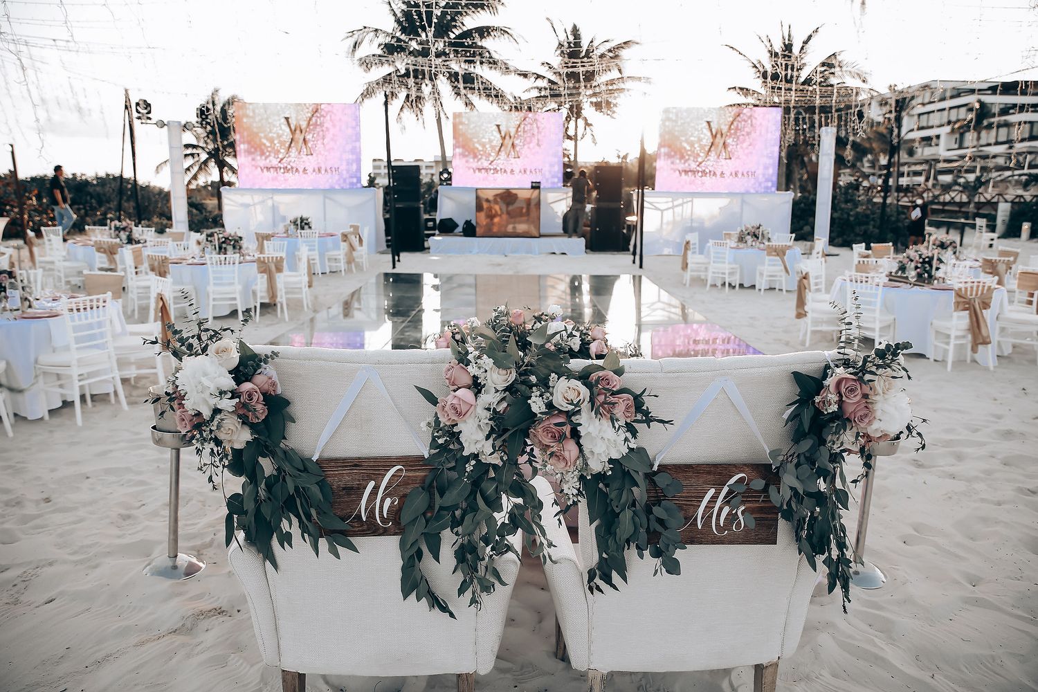 Elegant wedding reception setup with white tables, floral arrangements, and palm trees against a tropical beachfront setting.